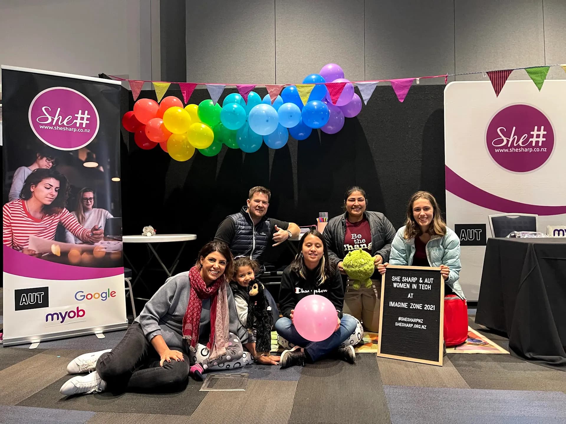 A group gathered on the floor in front of a stage decorated with rainbow balloons and bunting at the She Sharp Imagine Zone event.