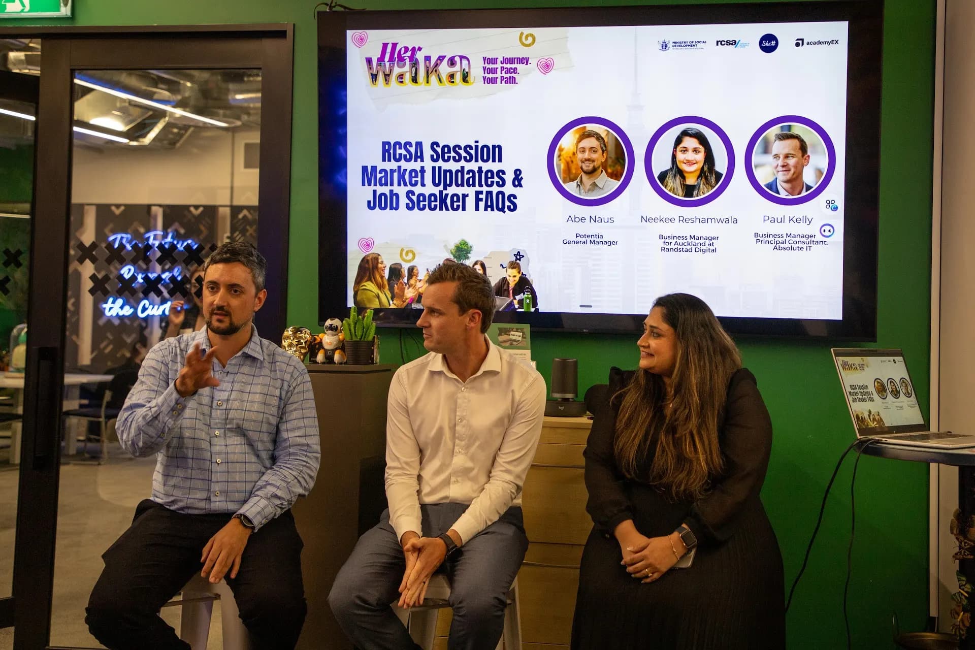 A panel of three speakers seated on stage during a She Sharp Her Waka session, with a presentation screen behind them.