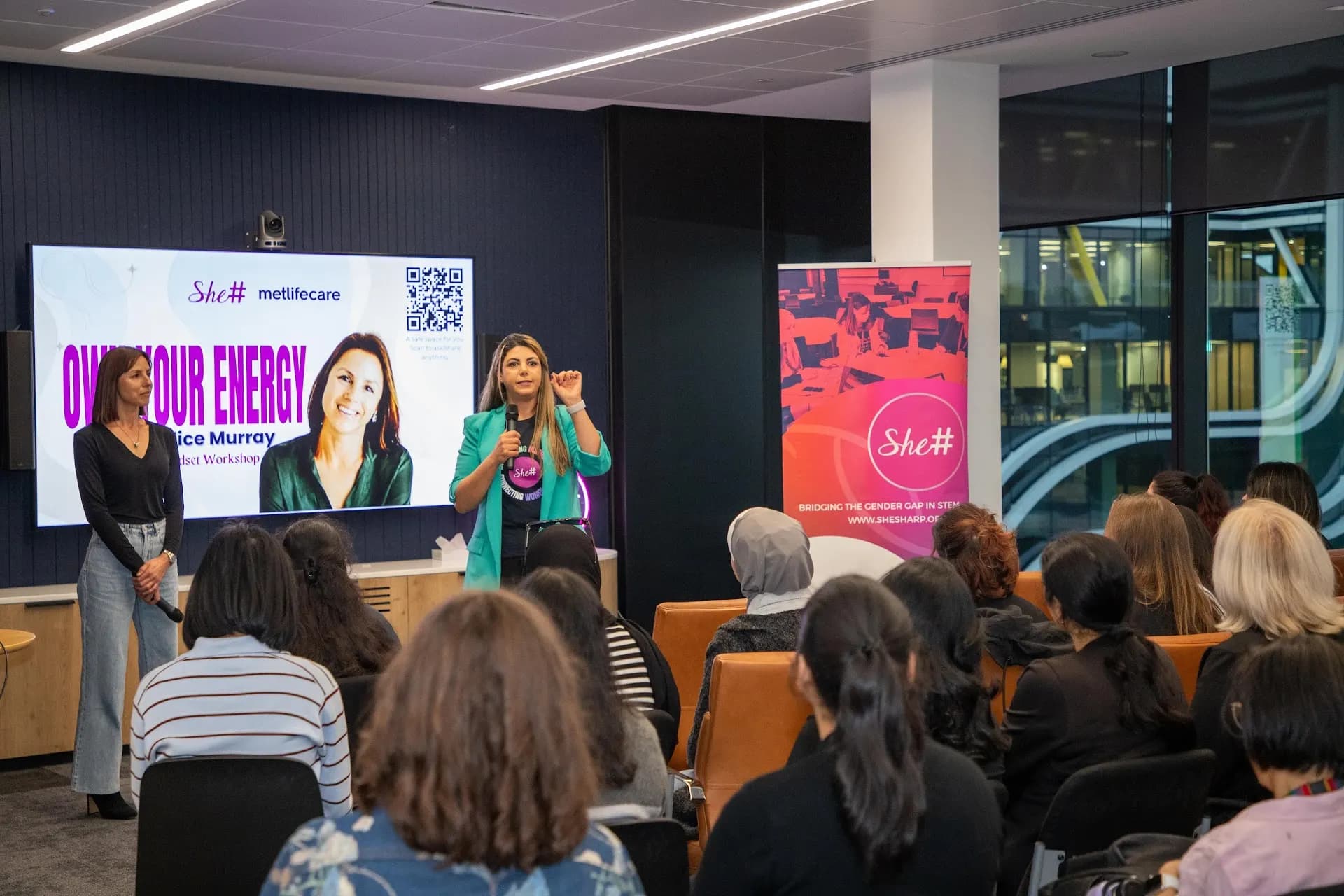 A presenter leading the Own Your Energy workshop at the front of the room before a seated She Sharp audience.