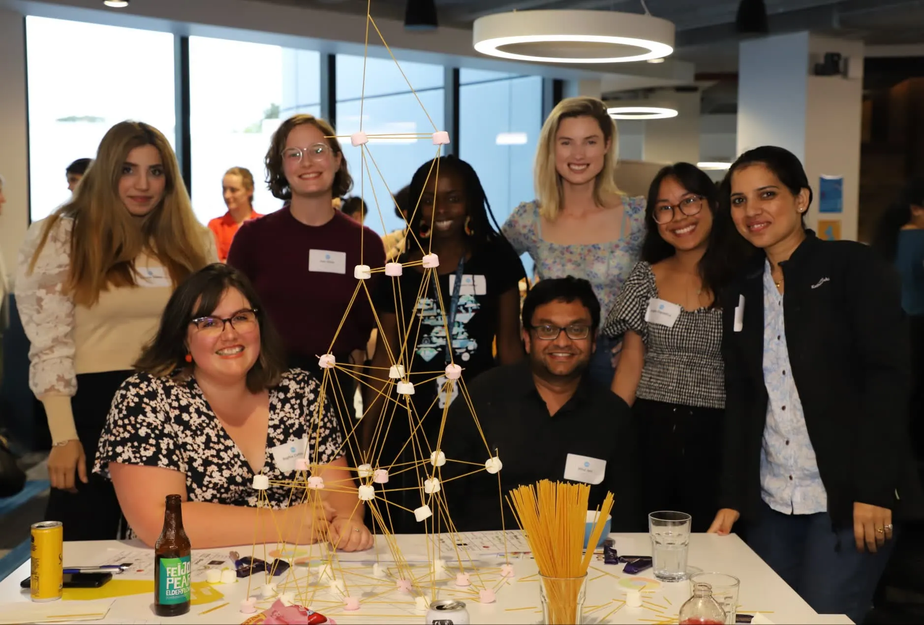 A diverse team smiling around a hands-on build challenge during a She Sharp workshop.