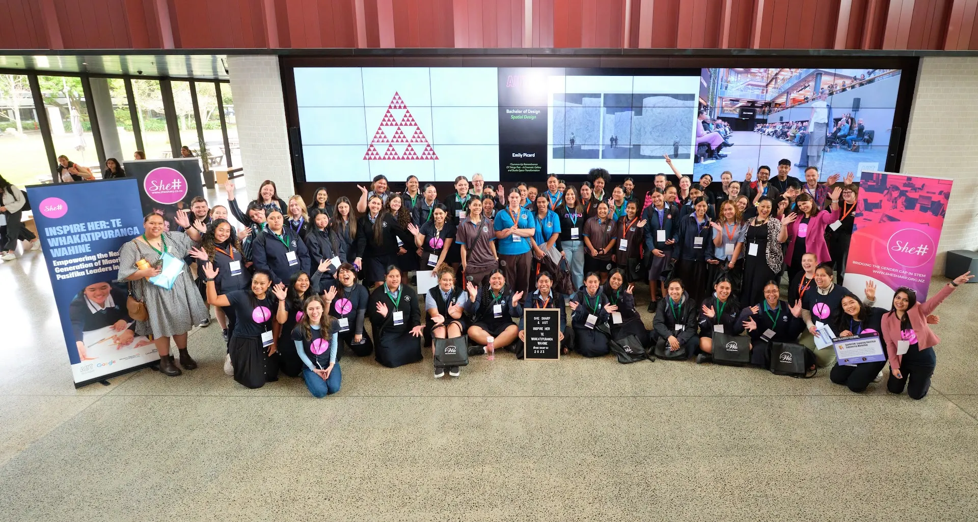A large gathering of the She Sharp community posed together in rows for a group photo at the Inspire Her event, many wearing pink lanyards.