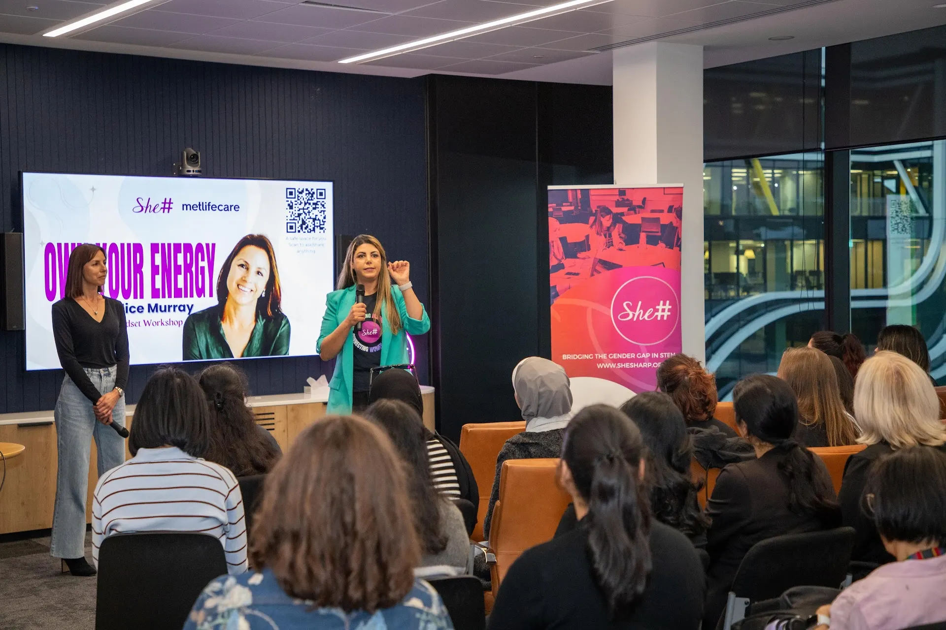 A presenter leading the Own Your Energy workshop at the front of the room before a seated She Sharp audience.